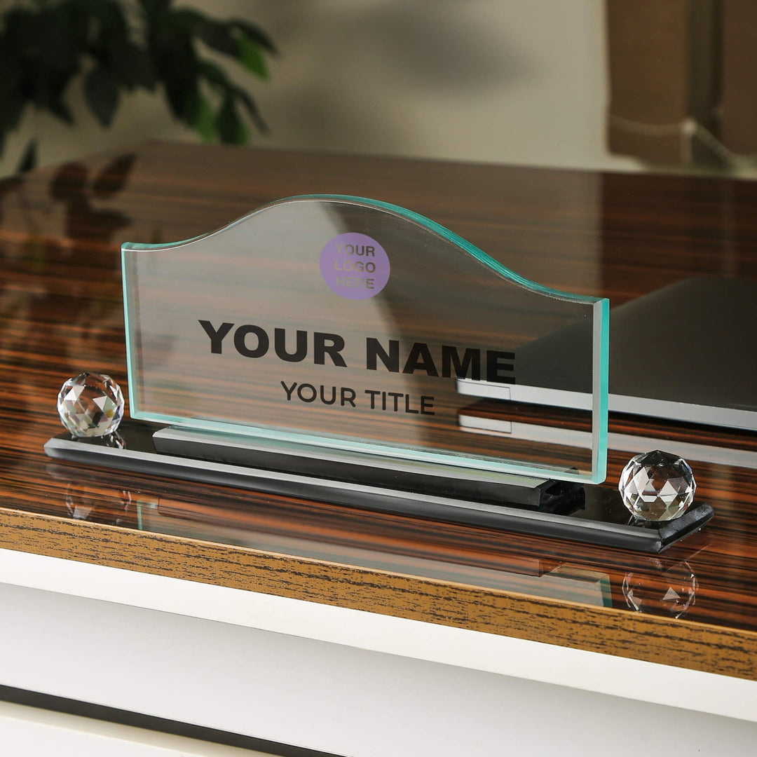 A crystal glass wave-shaped desk name plate with a placeholder for a name and title, displayed on a desk with the logo and name of a company above it.
