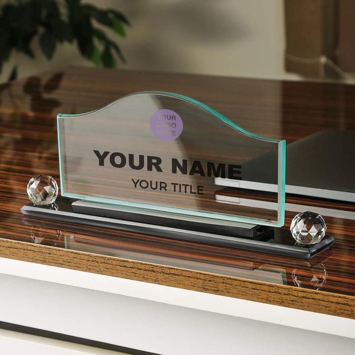 A crystal glass wave-shaped desk name plate with a placeholder for a name and title, displayed on a desk with the logo and name of a company above it.